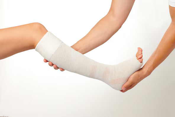 A close-up of a person's foot and lower leg wrapped in a light gray compression bandage, held by another person's hands against a plain white background.